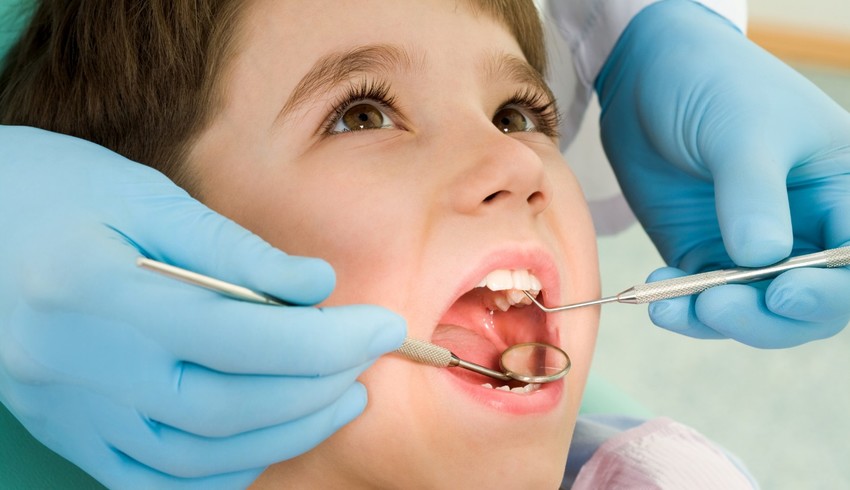 A imagem mostra um close-up de um procedimento odontológico, onde um profissional de saúde, usando luvas azuis, segura instrumentos dentários como um espelho bucal e uma sonda exploratória dentro da boca do paciente. A cena ocorre em um ambiente clínico e destaca a importância dos exames regulares para manutenção da saúde bucal.