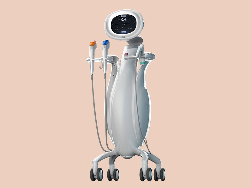 A imagem mostra um equipamento médico sobre rodas, projetado para uso clínico ou hospitalar. O dispositivo tem um design moderno e funcional, com uma tela na parte superior exibindo informações numéricas e textuais. Três instrumentos de mão com pontas coloridas (laranja, azul e verde) estão acoplados ao equipamento. O conjunto sugere um aparelho versátil, possivelmente utilizado para diferentes tipos de tratamentos ou diagnósticos médicos.